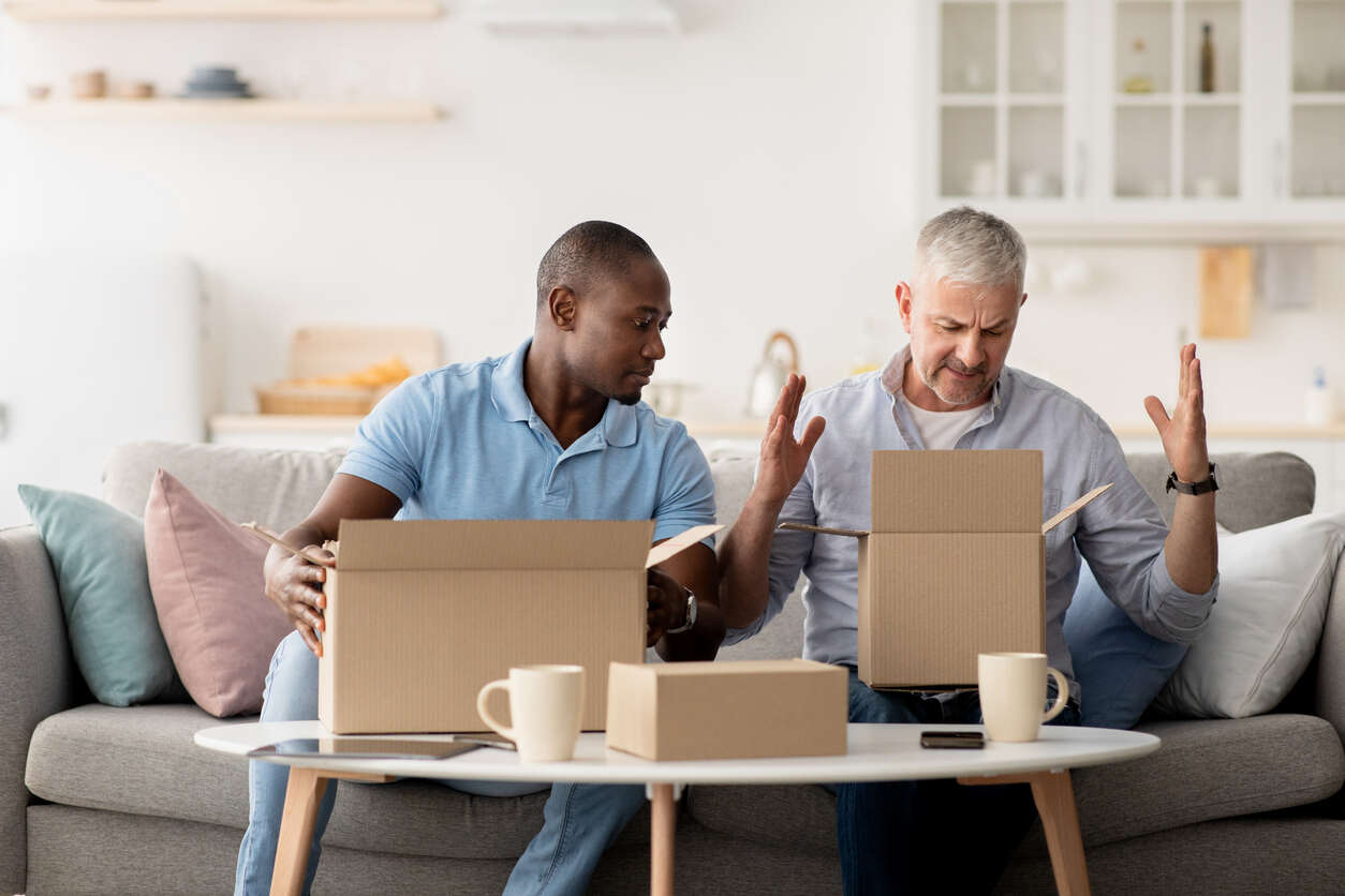 Men unboxing items on a coffee table look visibly upset and surprised, likely due to receiving defective household products.