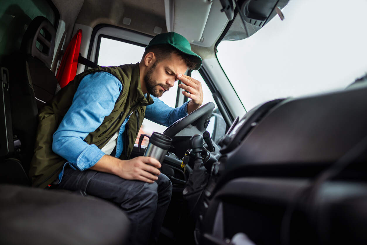  A fatigued truck driver sits in the cab of his truck with his hand on his forehead, representing the dangers of exhausted truckers who push past legal driving hours in Georgia