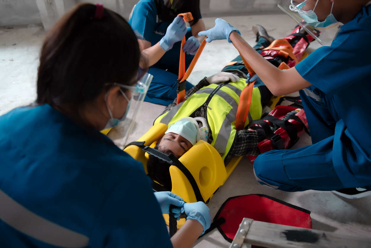 Medical personnel in face masks and gloves tending to an injured worker on a stretcher at an industrial site, symbolizing an industrial site injury.