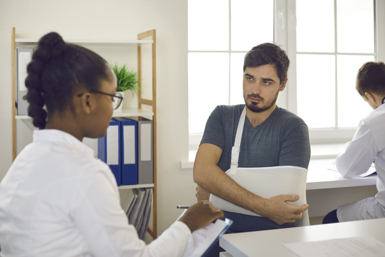 A serious male patient with his arm in a sling consults with a doctor, an American woman, highlighting the struggle for injury victims to get a proper diagnosis. The close-up shot of the doctor's office consultation visually represents the challenges faced by those seeking medical clarity after an injury