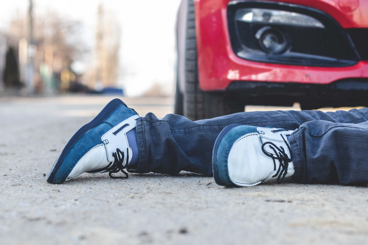 The legs and shoes of a person lying on the asphalt near the front of a red car, depicting the scene of a pedestrian collision in Marietta.