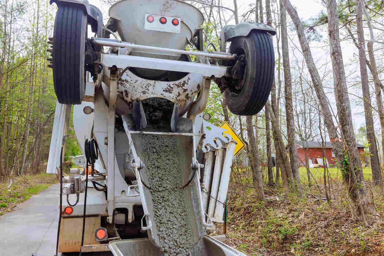  An up-close, eye-level shot of the back of a large concrete mixer truck. The truck is pulled over on a small, tree-lined road, with a home visible in the background. Which shows the concrete collision