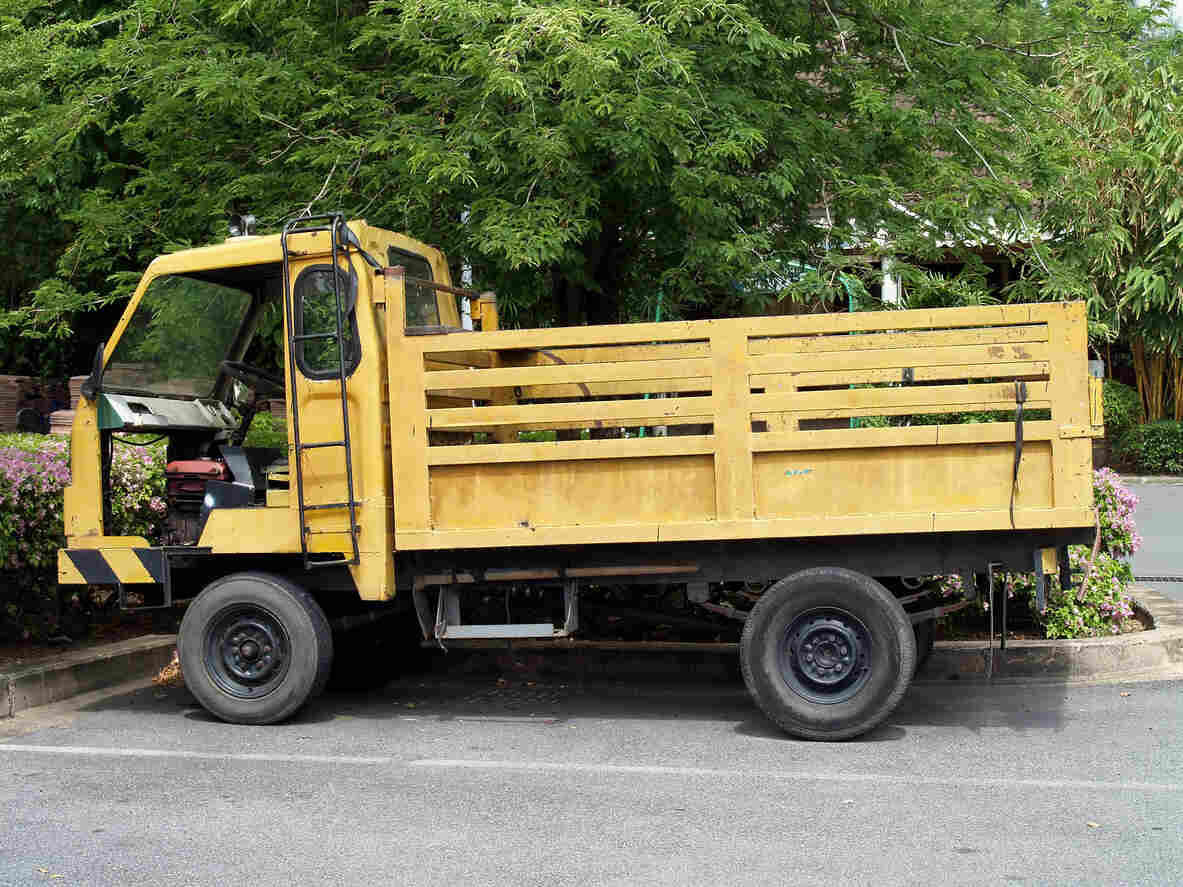 A small, yellow flatbed truck with wooden side panels is parked after met with an accident on an asphalt road next to a sidewalk with lush green trees and pink flowers in the background.