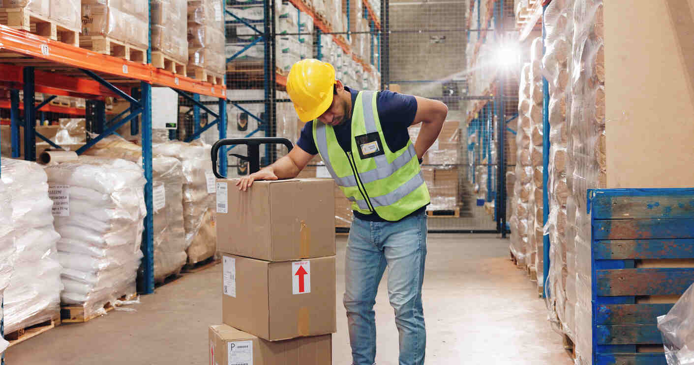 Warehouse worker in a reflective vest and hard hat experiencing back pain while handling boxes, representing workplace injury cases related to delivery companies like Amazon and FedEx.